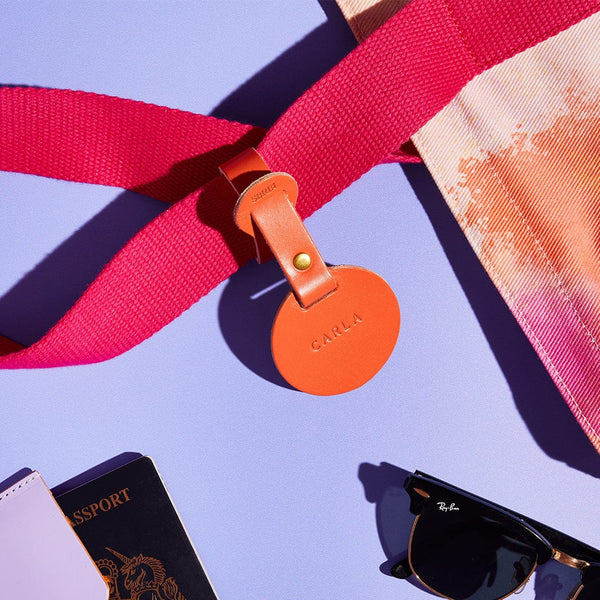An orange leather luggage tag personalised with the name 'Carla', surrounded by a beach bag, passport and sunglasses