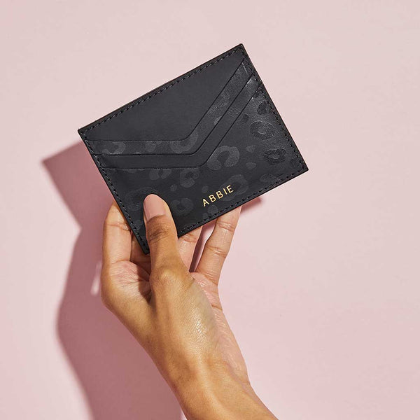 Black leather card holder with leopard print spots and personalised lettering, shown held against a pink background