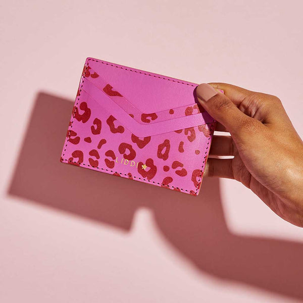 Fuchsia pink leather card holder with red leopard print spots and personalised lettering, shown held against a pink background
