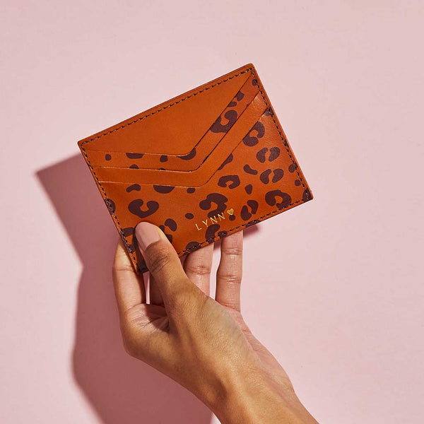 Tan brown leather card holder with leopard print spots and personalised lettering, shown held against a pink background